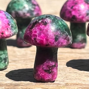 May include: Close-up of several mushroom-shaped sculptures. The sculptures have a dark purple stem and a cap that is a mix of pink and green. The mushrooms are made of a polished stone material.