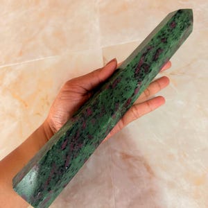 Può includere: Una torre di cristallo esagonale lucida con una base in zoisite verde screziata e inclusioni di rubino magenta scuro. La pietra presenta una superficie liscia e riflettente con una punta, mostrando motivi minerali naturali lungo la lunghezza.
