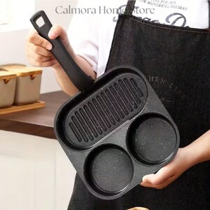 May include: A black non-stick frying pan with three cooking sections. The pan has a long, black handle and is designed for cooking multiple items at once. The pan has a textured surface and is held by a person.