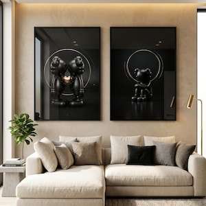 May include: Two framed wall art prints featuring black cartoon-style figures with X-shaped eyes. The left print shows two figures touching heads with a gold heart between them, while the right print shows a single figure sitting with knees pulled up.