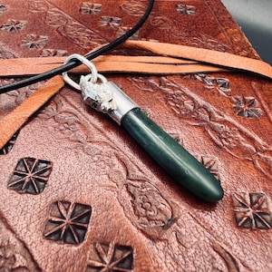 May include: A dark green jade pendant with a silver-colored metal cap and bail, suspended on a black cord. The pendant is displayed on a brown leather surface with embossed patterns.