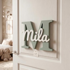 Könnte beinhalten: Ein salbeigrüner Holzbuchstabe "M" mit dem Namen "Mila" in weißer Schrift an einer weißen Tür befestigt. Ein kleines Etikett mit dem Text "ast" ist sichtbar. Die Tür ist leicht geöffnet und gibt einen Blick in ein Schlafzimmer frei.