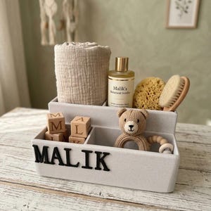 May include: A white organizer with the name "MALIK" in black letters, holding a rolled beige towel, a bottle of "Malik's Botanical Soothe", a natural sponge, a brush, wooden blocks, and a teddy bear teether.