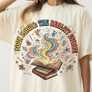 以下が含まれることがあります： クリーム色のTシャツ。開いた本と魔法の要素のカラフルなグラフィックが特徴です。「BOOK MAGIC: THE REALITY ESCAPE」の文字がデザインを囲むように配置されています。イラストには魔法使い、ドラゴン、その他のファンタジーのキャラクターが含まれています。