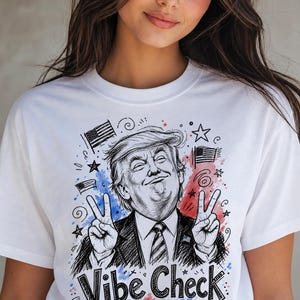 以下が含まれることがあります： 「Vibe Check」の文字と、ピースサインをする男性の漫画イラストが描かれた白いTシャツ。デザインには、アメリカの国旗、星、赤と青の水彩アクセントが含まれています。