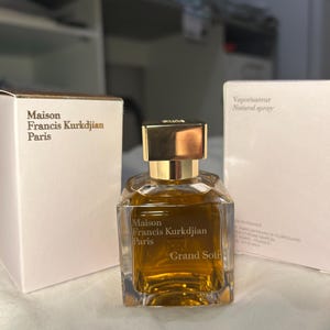 Peut inclure: Un flacon de parfum avec un bouchon doré et un corps en verre transparent de forme géométrique. Le flacon contient un liquide brun doré et porte l'inscription "Maison Francis Kurkdjian Paris Grand Soir". Deux boîtes blanches avec du texte doré sont de chaque côté.