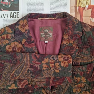 Pode incluir: Um casaco acolchoado com um padrão floral e paisley em tons de verde escuro, vermelho e dourado. O interior é forrado com tecido malva liso. Uma etiqueta no interior indica Ten Yards by Colmar com o tamanho 44. O casaco tem gola de lapela.