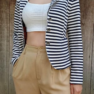 May include: A navy blue and white horizontal striped blazer with three buttons, a notched lapel, and two front pockets. The blazer is worn over a white cropped top with a textured pattern and paired with high-waisted tan trousers with two buttons.