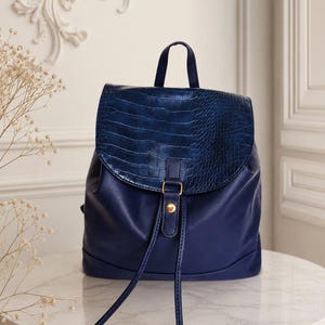 Puede incluir: Una mochila de color azul profundo con un acabado liso similar al cuero en el cuerpo principal y un patrón texturizado de cocodrilo en la solapa superior. La bolsa incluye una correa central vertical con un cierre de presión en tono dorado.
