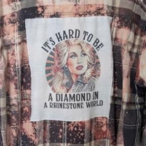 May include: A distressed flannel shirt with a faded plaid pattern in shades of black, gray, and peach. The back features a rectangular patch with a portrait of a woman and the text "IT'S HARD TO BE A DIAMOND IN A RHINESTONE WORLD."