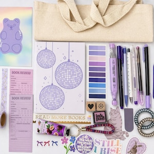 May include: A collection of purple and lavender stationery items, including pens, notebooks, stickers, and washi tape, arranged with a beige tote bag. The items have a disco ball and gummy bear theme. There is also a tube of passion fruit hand cream.