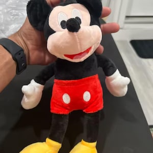 May include: A plush Mickey Mouse toy. The toy has a black body, ears, and arms, with white gloves. It wears red shorts with two white buttons and yellow shoes. The face is pink with black eyes and a red smile.