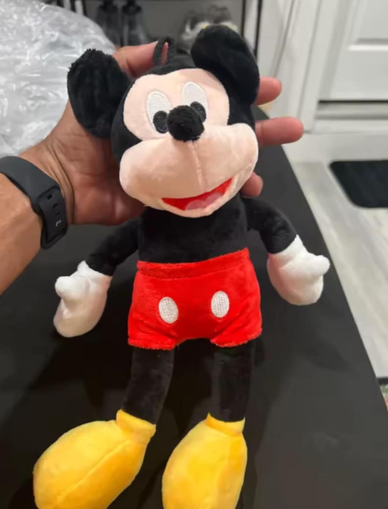 May include: A plush Mickey Mouse toy. The toy has a black body, ears, and arms, with white gloves. It wears red shorts with two white buttons and yellow shoes. The face is pink with black eyes and a red smile.