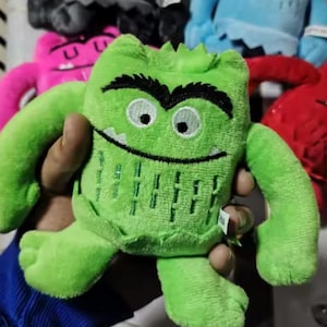 May include: A green plush monster toy with black eyebrows, a stitched smile, and white eyes. The toy has arms, legs, and a textured belly. Other colourful monster toys are visible in the background.