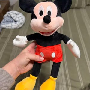 May include: A plush Mickey Mouse doll with black ears, a black body, red shorts with white buttons, yellow shoes, and white gloves. The doll has a pink face and a black loop for hanging.
