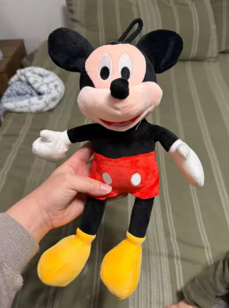 May include: A plush Mickey Mouse doll with black ears, a black body, red shorts with white buttons, yellow shoes, and white gloves. The doll has a pink face and a black loop for hanging.