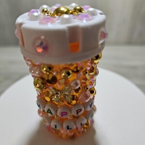 Puede incluir: Un frasco de pastillas decorativo, adornado con pedrer&iacute;a iridiscente, perlas y detalles dorados. El frasco presenta las palabras "HAPPY PILLS" deletreadas con cuentas de letras blancas. La parte superior es blanca y el cuerpo es una gradaci&oacute;n de naranja y dorado.