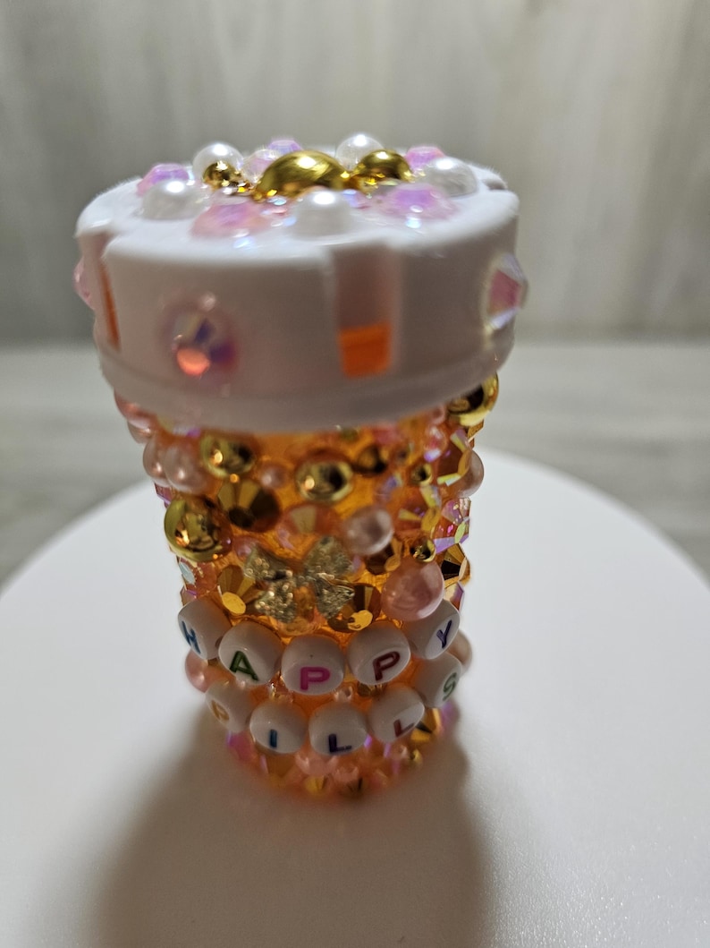 Puede incluir: Un frasco de pastillas decorativo, adornado con pedrer&iacute;a iridiscente, perlas y detalles dorados. El frasco presenta las palabras "HAPPY PILLS" deletreadas con cuentas de letras blancas. La parte superior es blanca y el cuerpo es una gradaci&oacute;n de naranja y dorado.