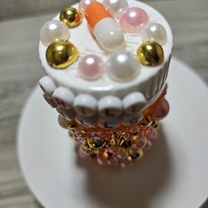 Puede incluir: Un objeto decorativo con tem&aacute;tica de pastillas. La parte superior es blanca con una pastilla naranja y blanca, cuentas doradas y perladas. La secci&oacute;n media tiene la palabra "PILLS" escrita con cuentas blancas. La base es un dise&ntilde;o de cuentas rosas y doradas. El objeto est&aacute; sobre un plato blanco.