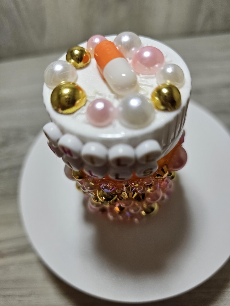 Puede incluir: Un objeto decorativo con tem&aacute;tica de pastillas. La parte superior es blanca con una pastilla naranja y blanca, cuentas doradas y perladas. La secci&oacute;n media tiene la palabra "PILLS" escrita con cuentas blancas. La base es un dise&ntilde;o de cuentas rosas y doradas. El objeto est&aacute; sobre un plato blanco.