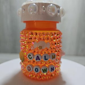 Puede incluir: Un frasco de pastillas naranja decorado con pedrer&iacute;a iridiscente y las palabras "CALM DOWN" deletreadas con cuentas de letras. La tapa del frasco es blanca y est&aacute; adornada con adornos transparentes en forma de burbujas.