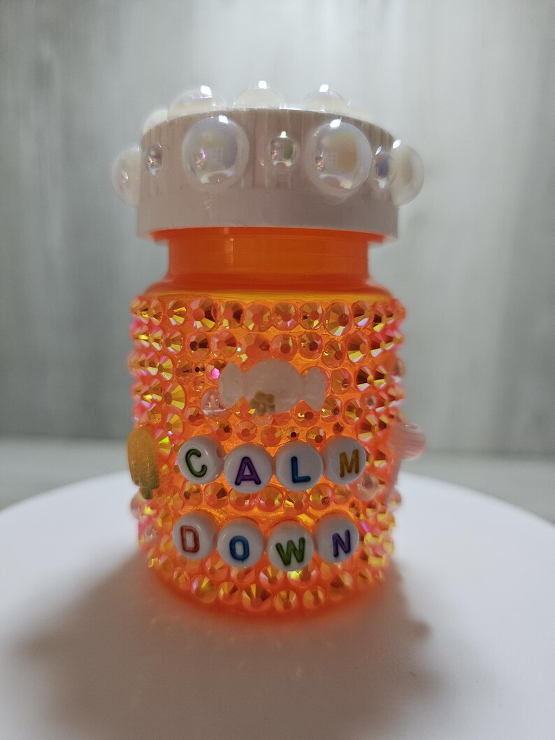Puede incluir: Un frasco de pastillas naranja decorado con pedrer&iacute;a iridiscente y las palabras "CALM DOWN" deletreadas con cuentas de letras. La tapa del frasco es blanca y est&aacute; adornada con adornos transparentes en forma de burbujas.