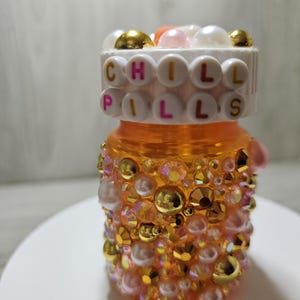 Puede incluir: Un frasco de pastillas naranja decorado con pedrer&iacute;a y perlas doradas, rosas e iridiscentes. La tapa del frasco presenta las palabras "CHILL PILLS" deletreadas con cuentas de letras. El frasco est&aacute; sobre una superficie blanca.