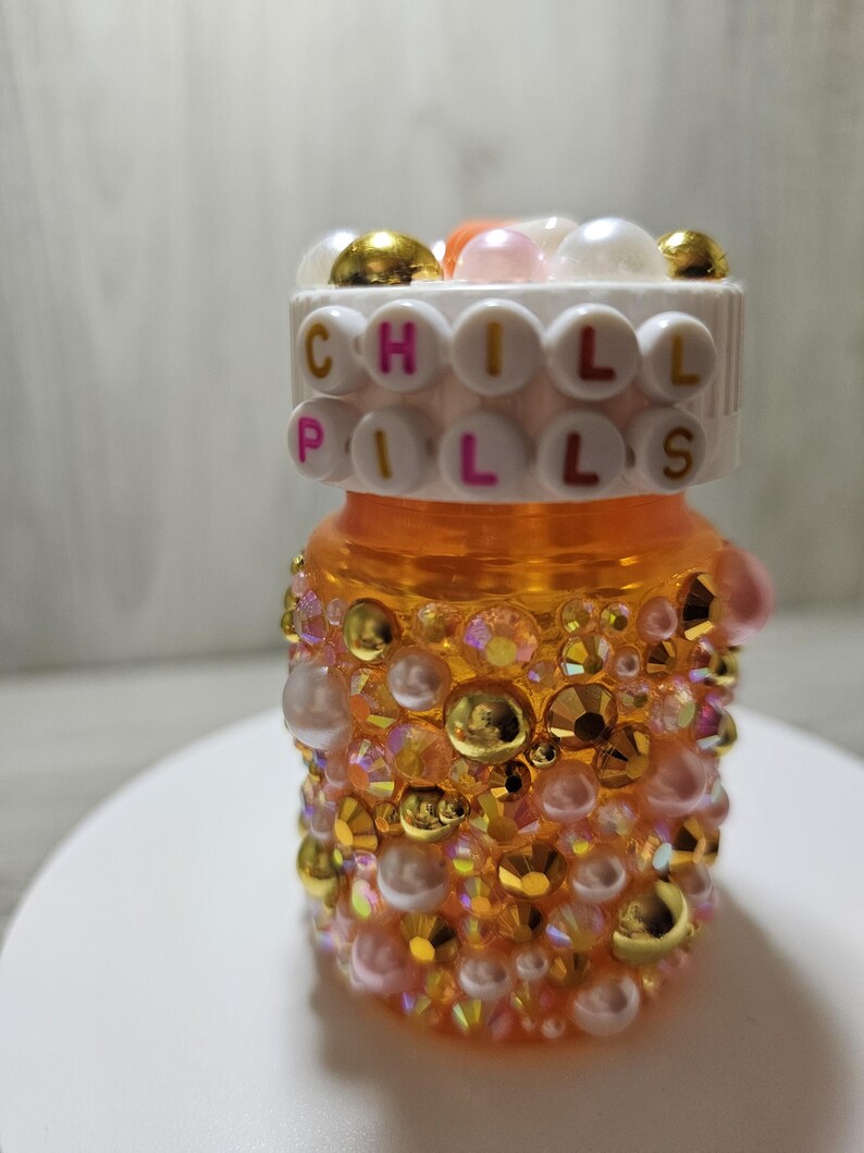 Puede incluir: Un frasco de pastillas naranja decorado con pedrer&iacute;a y perlas doradas, rosas e iridiscentes. La tapa del frasco presenta las palabras "CHILL PILLS" deletreadas con cuentas de letras. El frasco est&aacute; sobre una superficie blanca.