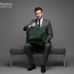 Pode incluir: Uma pasta de couro verde escuro com um bolso frontal e al&ccedil;as superiores. A pasta &eacute; segurada por uma pessoa que usa um terno cinza, camisa branca e sapatos pretos. Um acess&oacute;rio elegante para neg&oacute;cios.