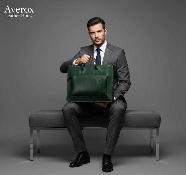 Pode incluir: Uma pasta de couro verde escuro com um bolso frontal e al&ccedil;as superiores. A pasta &eacute; segurada por uma pessoa que usa um terno cinza, camisa branca e sapatos pretos. Um acess&oacute;rio elegante para neg&oacute;cios.