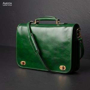 Pode incluir: Uma pasta de couro verde-escura com alça superior, alça de ombro removível e dois fechos giratórios de metal dourado na aba frontal. A bolsa exibe o texto Averox Leather House no canto superior esquerdo.