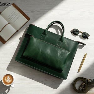 Pode incluir: Uma pasta de couro verde escuro com uma bolsa a condizer, al&ccedil;as e uma al&ccedil;a de ombro. A pasta est&aacute; sobre uma superf&iacute;cie de madeira branca, juntamente com um caderno, &oacute;culos de sol, uma caneta e uma ch&aacute;vena de caf&eacute;.