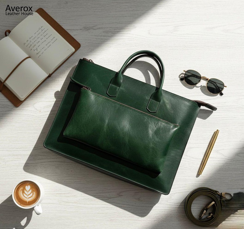 Pode incluir: Uma pasta de couro verde escuro com uma bolsa a condizer, al&ccedil;as e uma al&ccedil;a de ombro. A pasta est&aacute; sobre uma superf&iacute;cie de madeira branca, juntamente com um caderno, &oacute;culos de sol, uma caneta e uma ch&aacute;vena de caf&eacute;.