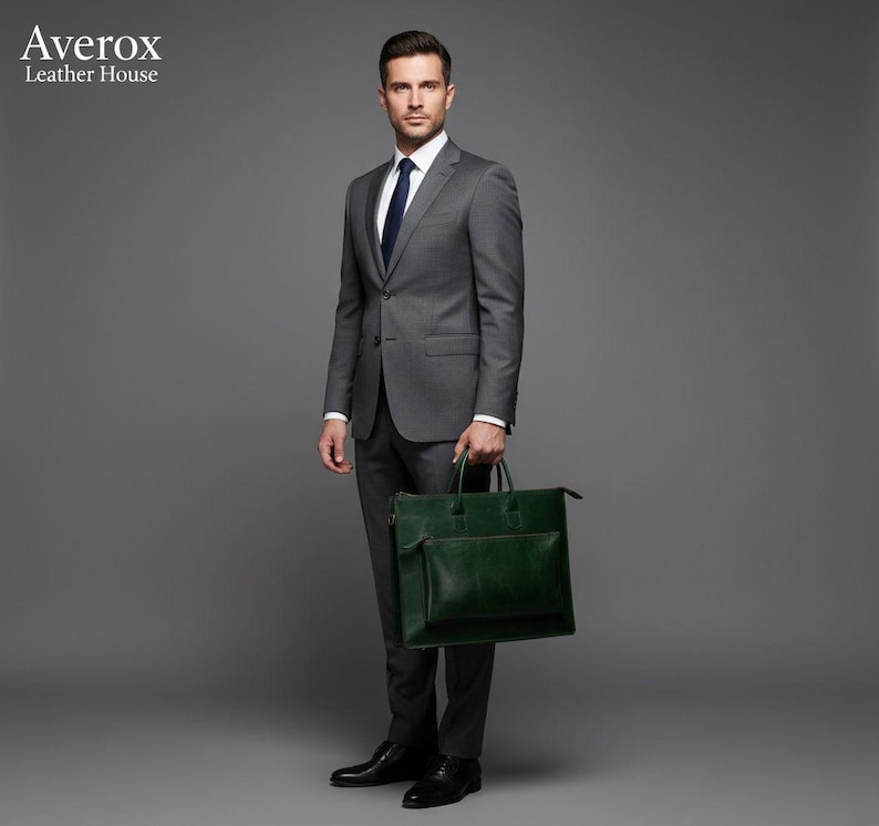 May include: A man in a grey suit and a navy blue tie holds a dark green leather briefcase. The briefcase has a front pocket and a zip closure. The image is set against a grey background.