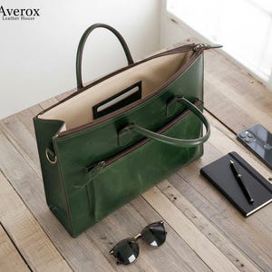 Pode incluir: Uma pasta de couro verde com uma al&ccedil;a superior e um bolso frontal com z&iacute;per. A bolsa est&aacute; aberta, revelando um interior bege. &Oacute;culos de sol, um caderno e uma caneta est&atilde;o em uma superf&iacute;cie de madeira.