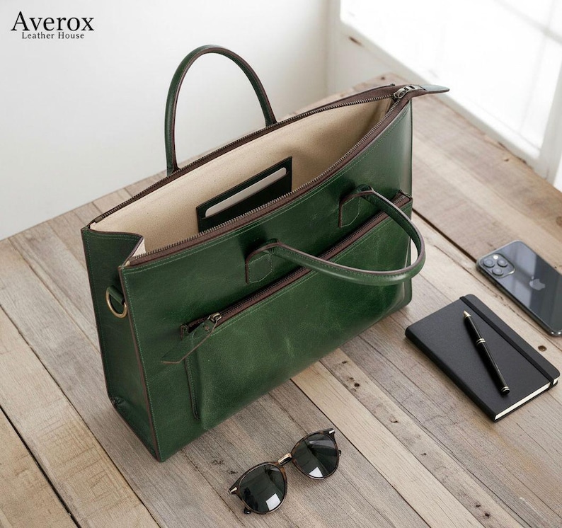 Pode incluir: Uma pasta de couro verde com uma al&ccedil;a superior e um bolso frontal com z&iacute;per. A bolsa est&aacute; aberta, revelando um interior bege. &Oacute;culos de sol, um caderno e uma caneta est&atilde;o em uma superf&iacute;cie de madeira.