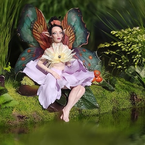 May include: A fantasy fairy doll with butterfly wings, a flower-like top, and a lavender petal skirt, seated near a pond. The wings are a mix of teal, orange, and brown. The doll has a delicate face and is surrounded by greenery.
