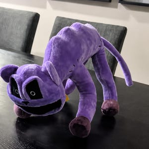Può includere: Un giocattolo di peluche a forma di gatto viola, con dettagli cuciti e accenti scuri. Il gatto ha un ampio sorriso e grandi occhi neri. Le zampe e le orecchie sono di un viola più scuro.