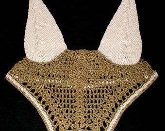 May include: A tan and white crochet horse bonnet with ear covers. The bonnet features a detailed openwork design in tan, with white ear covers and a white border along the bottom edge. The background is black.
