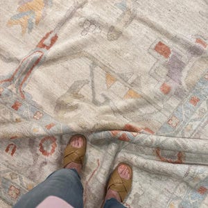 May include: An overhead shot of a beige rug with a detailed pattern in shades of blue, orange, and red. The rug is partially folded, revealing a glimpse of the underside. Tan leather sandals and blue jeans are visible.