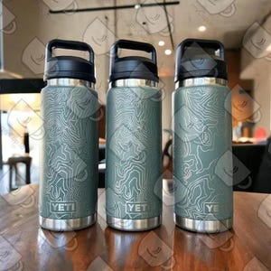 May include: Three sage green YETI water bottles with black lids and handles. Each bottle features a white topographic map line pattern etched onto the exterior surface and the YETI brand logo printed near the base of the metal body.
