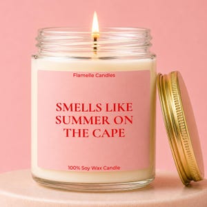 May include: A clear glass jar candle with a lit wick, filled with white wax. The label is light pink with red text reading Flamelle Candles, SMELLS LIKE SUMMER ON THE CAPE, and 100% Soy Wax Candle. A gold metal lid sits beside the jar.