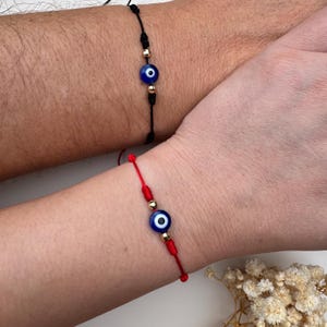 May include: Two adjustable cord bracelets, one black and one red, each featuring a central blue and white evil eye bead flanked by two small metallic gold-toned beads. The bracelets are designed with a simple sliding knot closure mechanism.