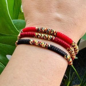 May include: Three stacked bracelets made of braided cord. The top bracelet is solid red, the middle is deep red, and the bottom is black. Each bracelet features a section of multicoloured woven thread in yellow, green, and red patterns.