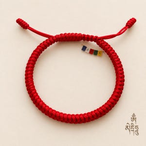 May include: A bright red braided cord bracelet featuring a sliding knot closure with two dangling ends. Attached to the bracelet is a small, miniature string of five colourful rectangular prayer flags in blue, white, red, green, and yellow.