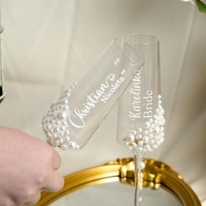 May include: Two clear glass champagne flutes decorated with white pearls at the base. One flute features the white printed names Christian and Nicoleta, while the other flute displays the white printed names Karolinka and Bride in a script font.