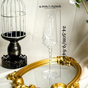May include: A clear glass champagne flute decorated with white pearls on the bowl and stem. The glass features the engraved names Christian and Nicoleta. It measures 1.65 inches in diameter and 9.64 inches in height as indicated by text.