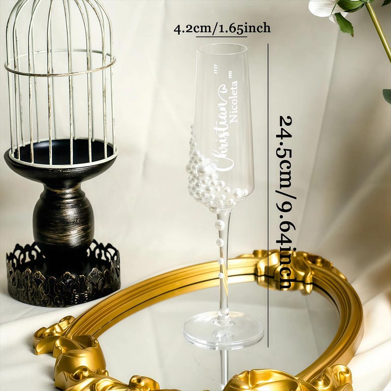 May include: A clear glass champagne flute decorated with white pearls on the bowl and stem. The glass features the engraved names Christian and Nicoleta. It measures 1.65 inches in diameter and 9.64 inches in height as indicated by text.