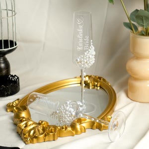 May include: Two clear glass champagne flutes decorated with white pearls. One flute is upright and engraved with the text "Karolinka Bride" and a heart. The other flute lies horizontally and is engraved with the text "Christian Nicoleta" and a heart.