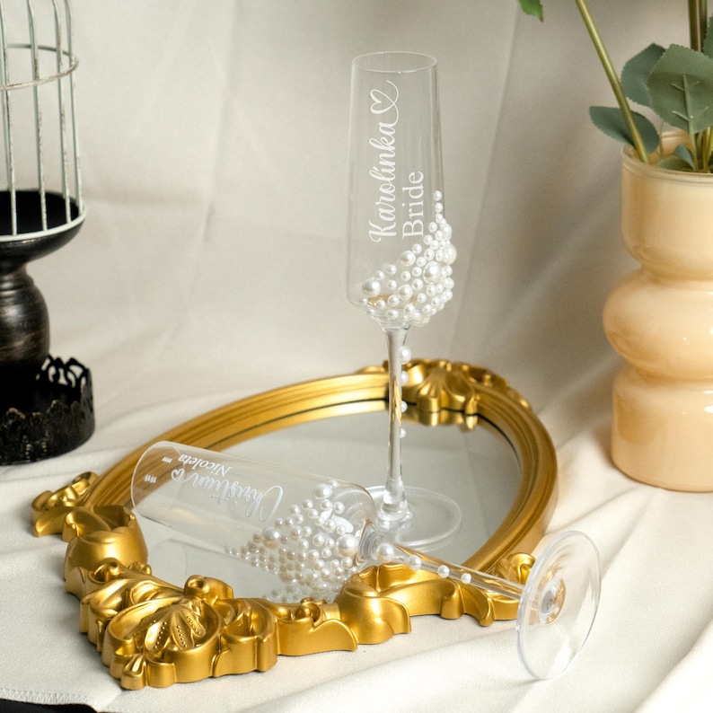 May include: Two clear glass champagne flutes decorated with white pearls. One flute is upright and engraved with the text "Karolinka Bride" and a heart. The other flute lies horizontally and is engraved with the text "Christian Nicoleta" and a heart.